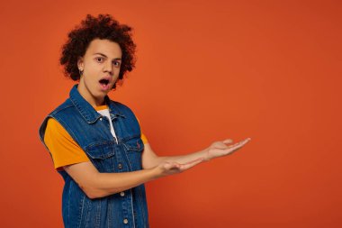 astonished emotional african american man in urban outfit gesturing actively on orange backdrop clipart