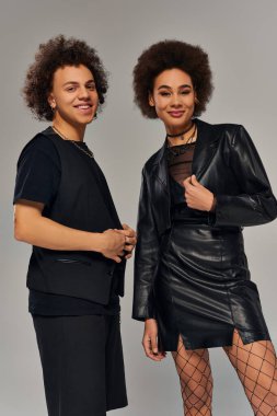 Gri zeminde birlikte poz veren şık siyah şehir kıyafetleri içindeki neşeli Afro-Amerikan kardeşler.