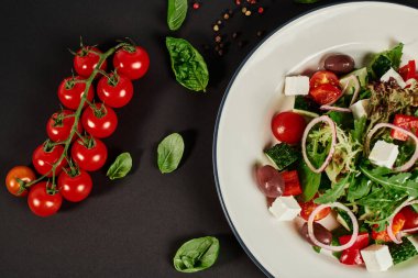 Siyah arkaplanda vişneli domateslerin yanında geleneksel Yunan salatasıyla tabağın üst görüntüsü