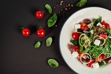 Siyah arkaplanda vişneli domateslerin yanında geleneksel Yunan salatasıyla tabağın üst görüntüsü