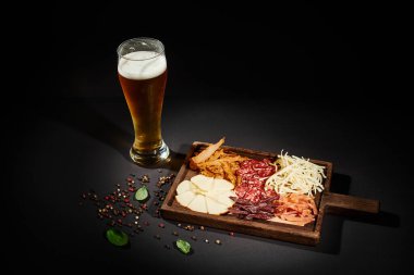 glass of cold beer near charcuterie board with gourmet cheese, dried beef and salami on black clipart