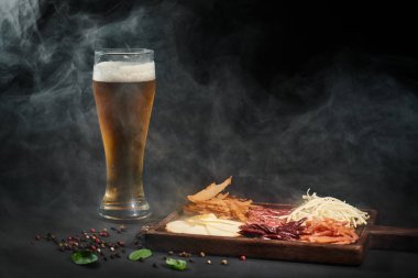 Şarküteri masasının yanında bir bardak bira gurme peyniri, kurutulmuş biftek ve siyah arka planda salam.