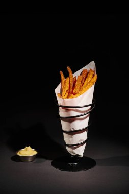 homemade and salty French fries inside of paper cone near dipping sauce on wooden cutting board clipart