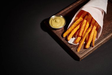 delicious homemade French fries inside of paper cone near dipping sauce on wooden cutting board clipart