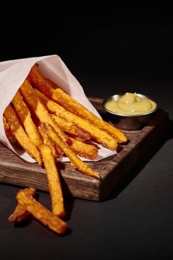 vertical shot of salty French fries inside of paper cone near dipping sauce on wooden cutting board clipart