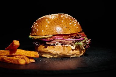 Lezzetli hamburger, kırmızı soğanlı, domatesli ve patates kızartmasının yanında siyah arka planda.