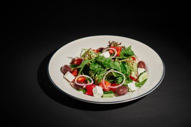 Hafif yemek, peynirli Yunan salatası, kırmızı soğan, siyah arkaplanda roka yaprakları.