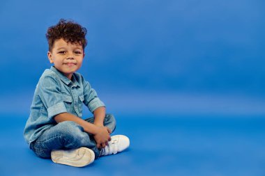 smiling and cute african american preschooler boy in stylish denim outfit sitting on blue background clipart