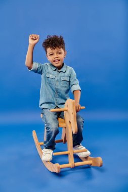 cute african american preschooler boy in stylish denim clothes sitting on rocking horse on blue clipart