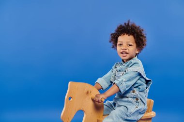 Mutlu Afro-Amerikan çocuk kot elbiseli sallanan sandalyede mavi arka planda oturuyor.