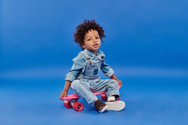 Mutlu Afro-Amerikan çocuk kot elbiseli, mavi arka planda panoda oturuyor.