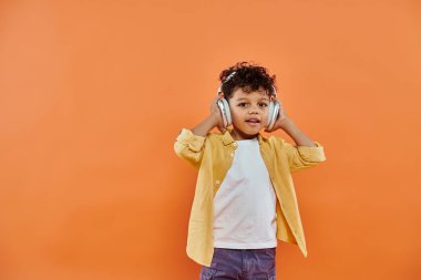 Mutlu ve kıvırcık Afrikalı Amerikalı çocuk turuncu arka planda kablosuz kulaklıkla müzik dinliyor.
