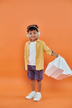 happy african american boy listening music in headphones and holding shopping bags on orange clipart