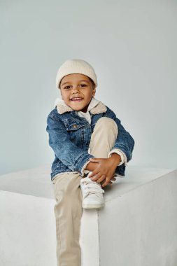 happy african american boy in winter attire and beanie hat sitting on concrete cube on grey clipart