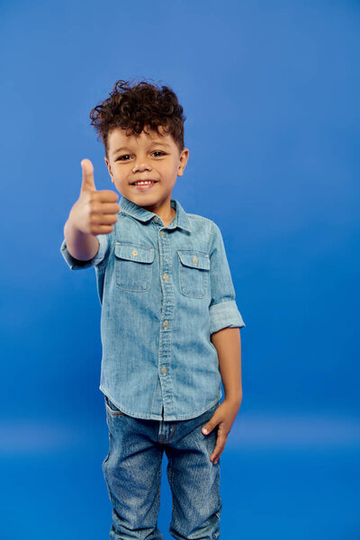 curly african american preschooler boy in stylish denim outfit showing like on blue background