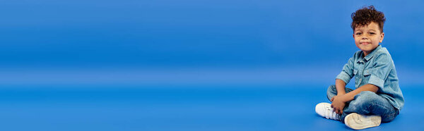 счастливый африканский американский дошкольник в стильном джинсовом наряде сидит на синем фоне, баннер