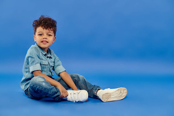 улыбающийся африканский американский дошкольник в стильной джинсовой одежде, сидящий на синем фоне