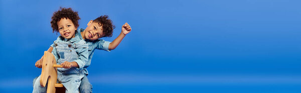 Симпатичные африканские американские братья в джинсовой одежде сидят вместе на качающейся лошади на голубом, баннер