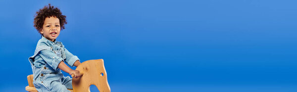 веселый африканский мальчик-малыш в стильной джинсовой одежде, сидящий на деревянном кресле-качалке, баннер