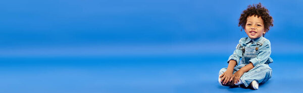 счастливый африканский американский мальчик в джинсовой одежде, сидящий со скрещенными ногами на синем баннере
