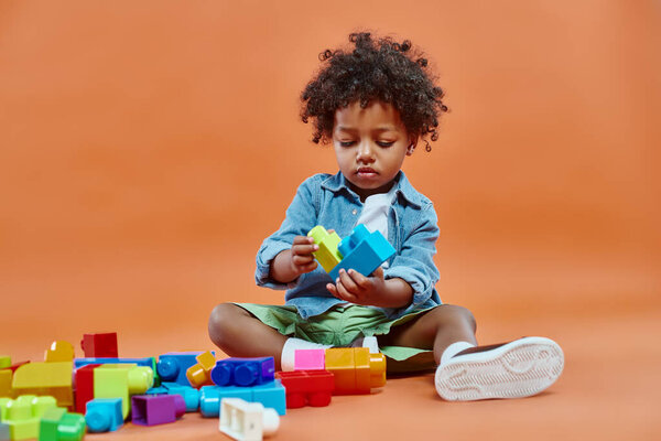 adorable african american toddler boy in casual attire sitting and playing building blocks on orange