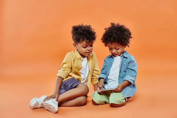 穿着休闲装的快乐的非洲裔美国孩子看着蹒跚学步的弟弟玩智能手机 — 图库照片