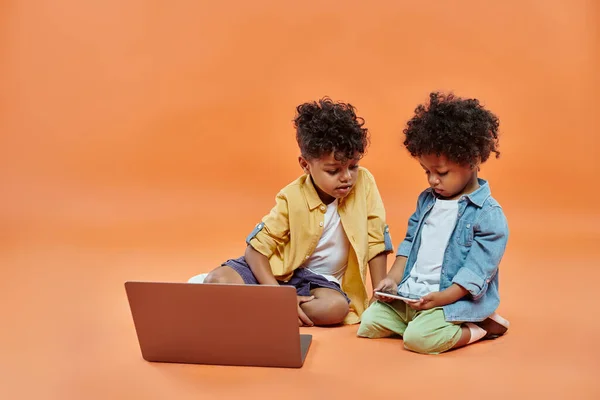 african american boy in casual attire looking at toddler brother playing on smartphone near laptop