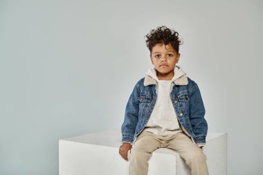 curly african american boy in winter attire and beanie sitting on concrete cube on grey backdrop clipart