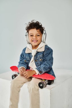 Mutlu Afro-Amerikalı kışın giyinmiş ve kulaklıklı çocuk küpün üzerinde gri pano ile oturuyor.