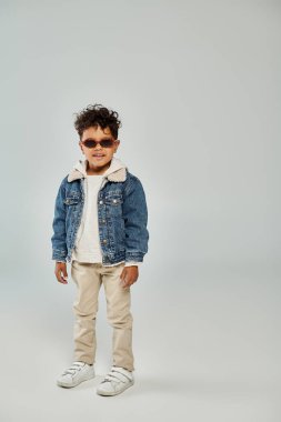 cheerful curly african american boy in winter attire and sunglasses standing on grey backdrop clipart