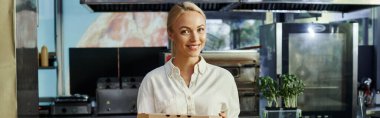 Elinde pizza kutusu olan neşeli sarışın menajer ve modern kafede kameraya gülümsüyor.
