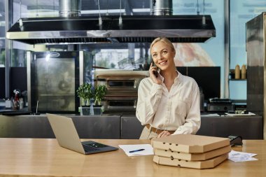 Akıllı telefondan konuşan neşeli sarışın kadın ve kafede pizza kutularının ve laptopun yanında sipariş kabul eden.