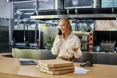 Pizza kutularının yanında akıllı telefondan konuşan heyecanlı sarışın kadın ve modern kafede tezgahta dizüstü bilgisayar.