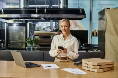 Pizza kutularının yanında akıllı telefondan mesajlaşan pozitif sarışın kadın ve modern kafede tezgahta dizüstü bilgisayar.