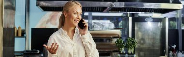 Heyecanlı sarışın yönetici cep telefonuyla konuşuyor ve modern kafede el kol hareketi yapıyor, yatay afiş.