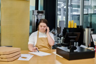 Down sendromlu genç bir kadın para terminali yakınında akıllı telefondan ve kafede pizza kutularından konuşuyor.