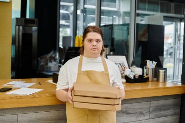 Kafede çalışırken elinde pizza kutuları olan ve kameraya bakan zihinsel engelli genç bir kadın.