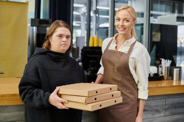Down sendromlu genç bir kadın çalışanın elinde kahve dükkanında gülümseyen bir yöneticinin yanında pizza kutuları var.