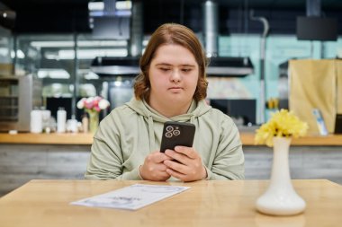 Down sendromlu genç bir kadın modern kafede menü kartının yanında akıllı telefondan sohbet ediyor.