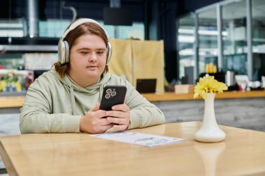 Down sendromlu bir kadın akıllı telefon tutuyor ve rahat bir kafede kulaklıkla müzik dinliyor.
