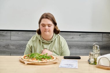 Down sendromlu genç bir kadın modern, rahat bir kafede otururken lezzetli pizzalara bakıyor.