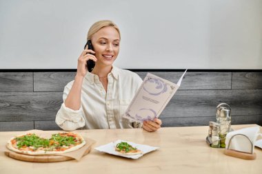 Akıllı telefondaki gülümseyen sarışın kadın ve kafedeki lezzetli pizzanın yanındaki menü kartına bakıyor.