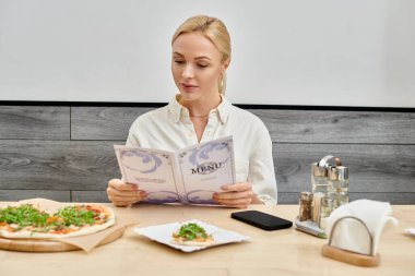 Modern kafede otururken lezzetli pizzanın yanında menüye bakan şık sarışın kadın.