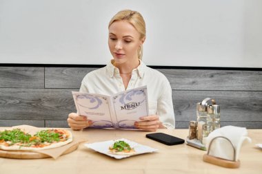 stylish blonde woman looking in menu card near delicious pizza while sitting in modern cafe clipart