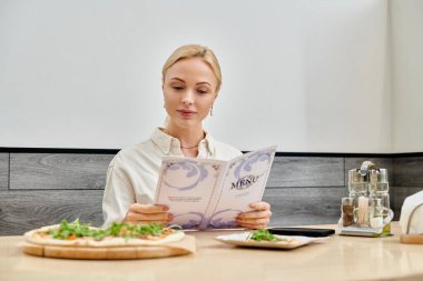 Güzel sarışın kadın modern kafede otururken lezzetli pizzanın yanındaki menüye bakıyor.
