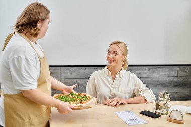 Down sendromlu genç garson modern kafede neşeli kadınlara lezzetli pizzalar öneriyor.