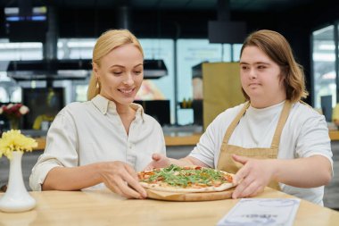 Down sendromlu genç garson modern kafede neşeli kadınlara lezzetli pizzalar öneriyor.