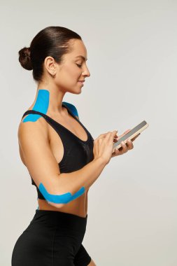 Siyah spor giymiş neşeli güzel bir kadın vücudunda kineziyolojik bantlarla telefonuna bakıyor.