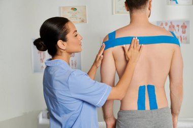Tıbbi kostümlü çekici kadın doktor randevu sırasında erkeğin üzerine kinezyolojik bantlar yapıştırıyor.