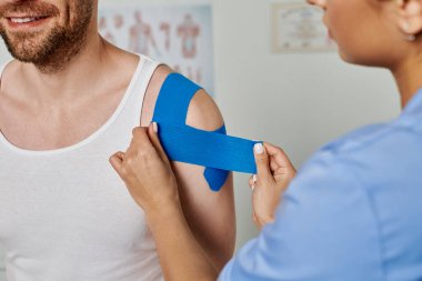 Kadın doktor vücuduna kineziyolojik bantlar yerleştirirken hastanın gülümseyişinin kırpılmış görüntüsü.